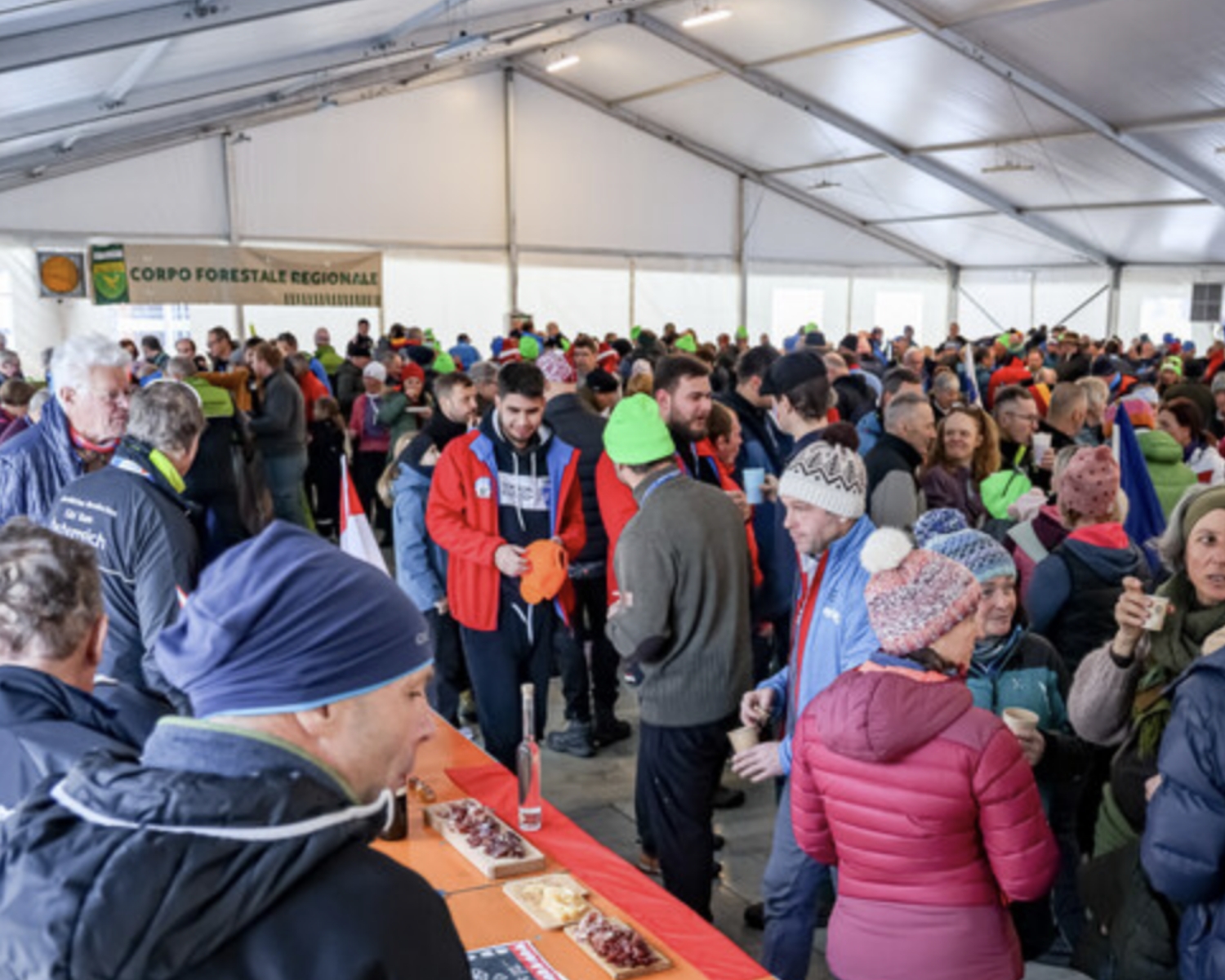 Una grande folla di persone, molte delle quali con abiti e cappelli invernali, si riunisce all'interno di una grande tenda. Alcuni stanno vicino a un tavolo con del cibo, mentre altri si mescolano. Sullo sfondo è appeso un cartello "Corpo Forestale Regionale".