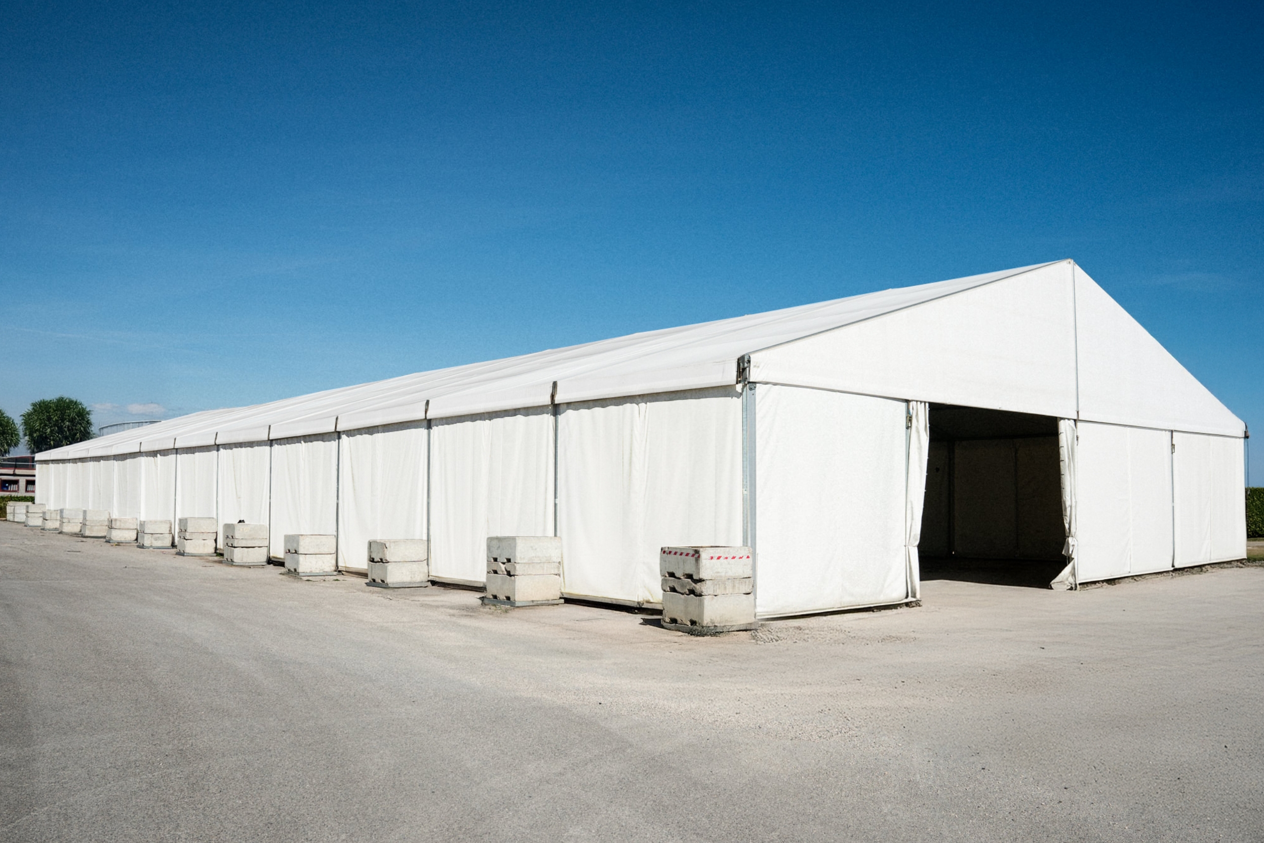 Un grande tendone bianco con pareti laterali e sezioni frontali aperte si trova su un'area pavimentata sotto un cielo azzurro e limpido, con blocchi di cemento che fissano i bordi del tendone.