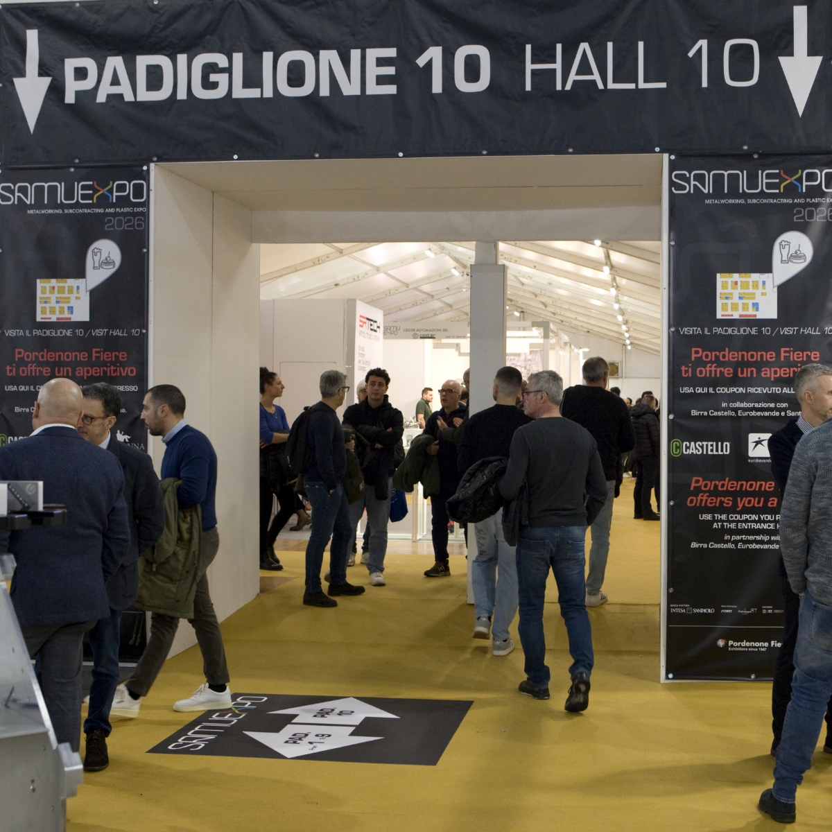 Le persone attraversano l'ingresso del Padiglione 10 al SamuExpo 2024. Il padiglione ha grandi insegne sopra e ai lati e il pavimento è giallo. L'evento appare affollato e vivace, con molti partecipanti.