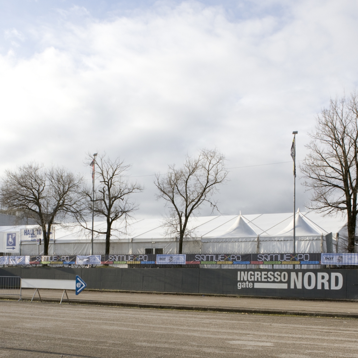 Un grande tendone bianco per eventi si trova dietro una recinzione con striscioni e cartelli. Sono visibili alberi senza foglie e un cielo nuvoloso. Un cartello sulla recinzione recita "INGRESSO gate NORD.
