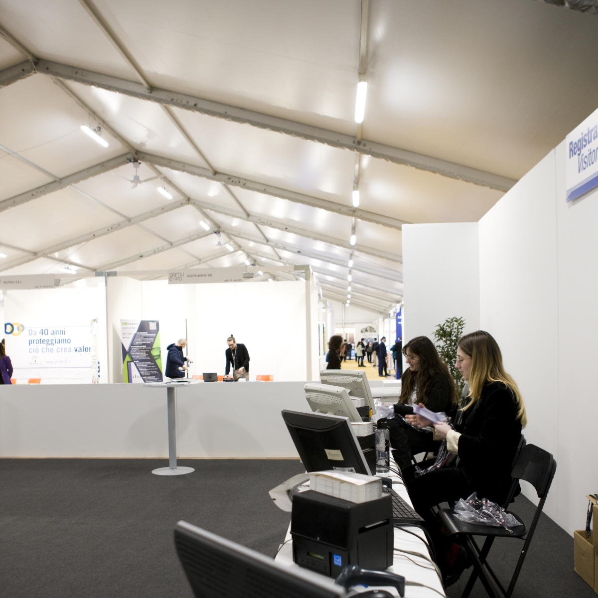 Una sala eventi spaziosa e ben illuminata con persone che camminano e due donne sedute al banco di registrazione con computer in primo piano. Pareti bianche e un alto soffitto a tenda circondano l'area.