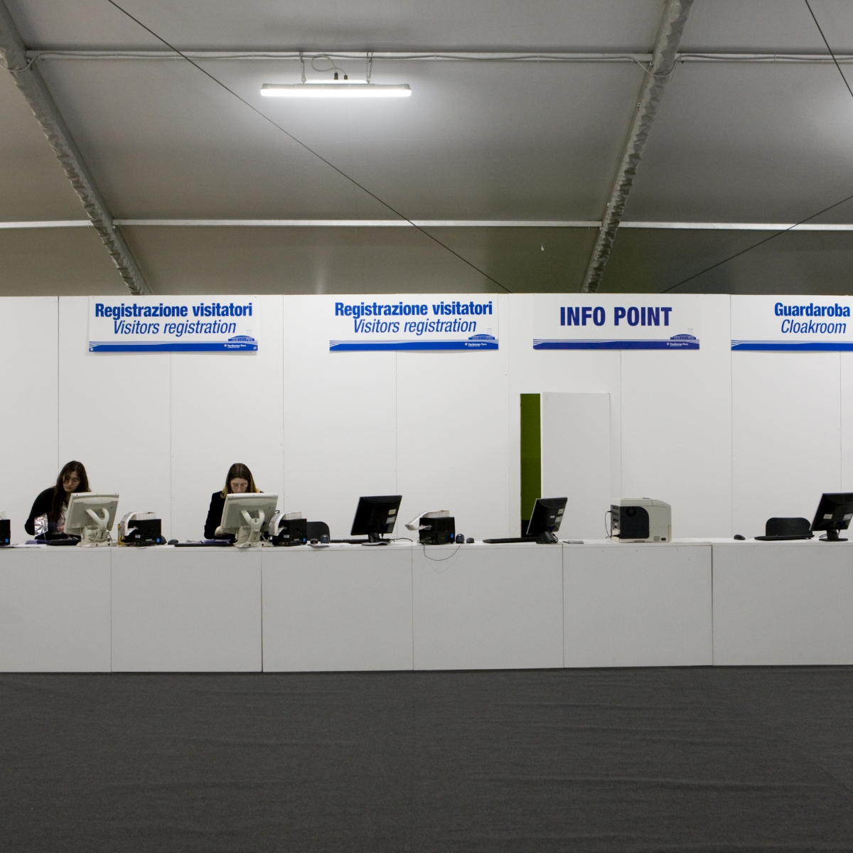 Un banco di registrazione e informazioni durante un evento, con due donne sedute dietro a dei computer. I cartelli in alto recitano "Registrazione visitatori", "INFO POINT" e "Guardaroba" in italiano e in inglese.