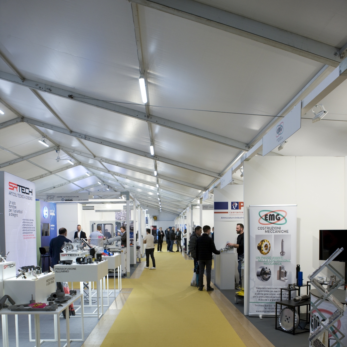 Sala espositiva di una fiera, con stand su entrambi i lati di un corridoio di moquette gialla, che espongono prodotti industriali. Diverse persone camminano, curiosano e parlano in vari stand.