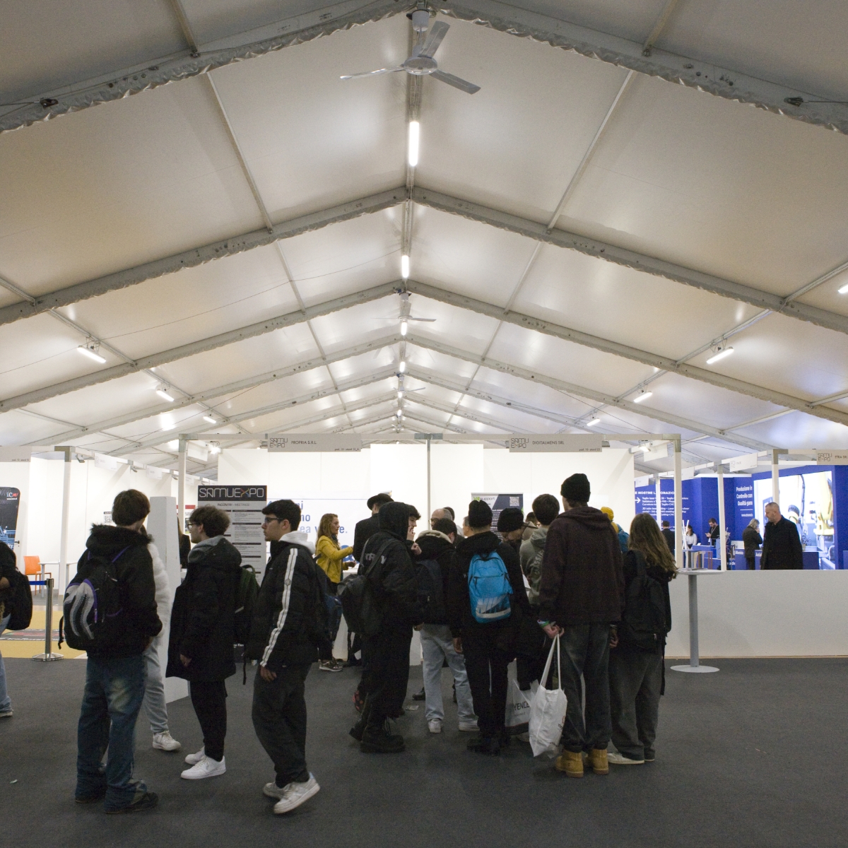 Un gruppo di persone con zaini e borse è in fila all'interno di un grande tendone bianco, con stand informativi e personale visibili sullo sfondo.