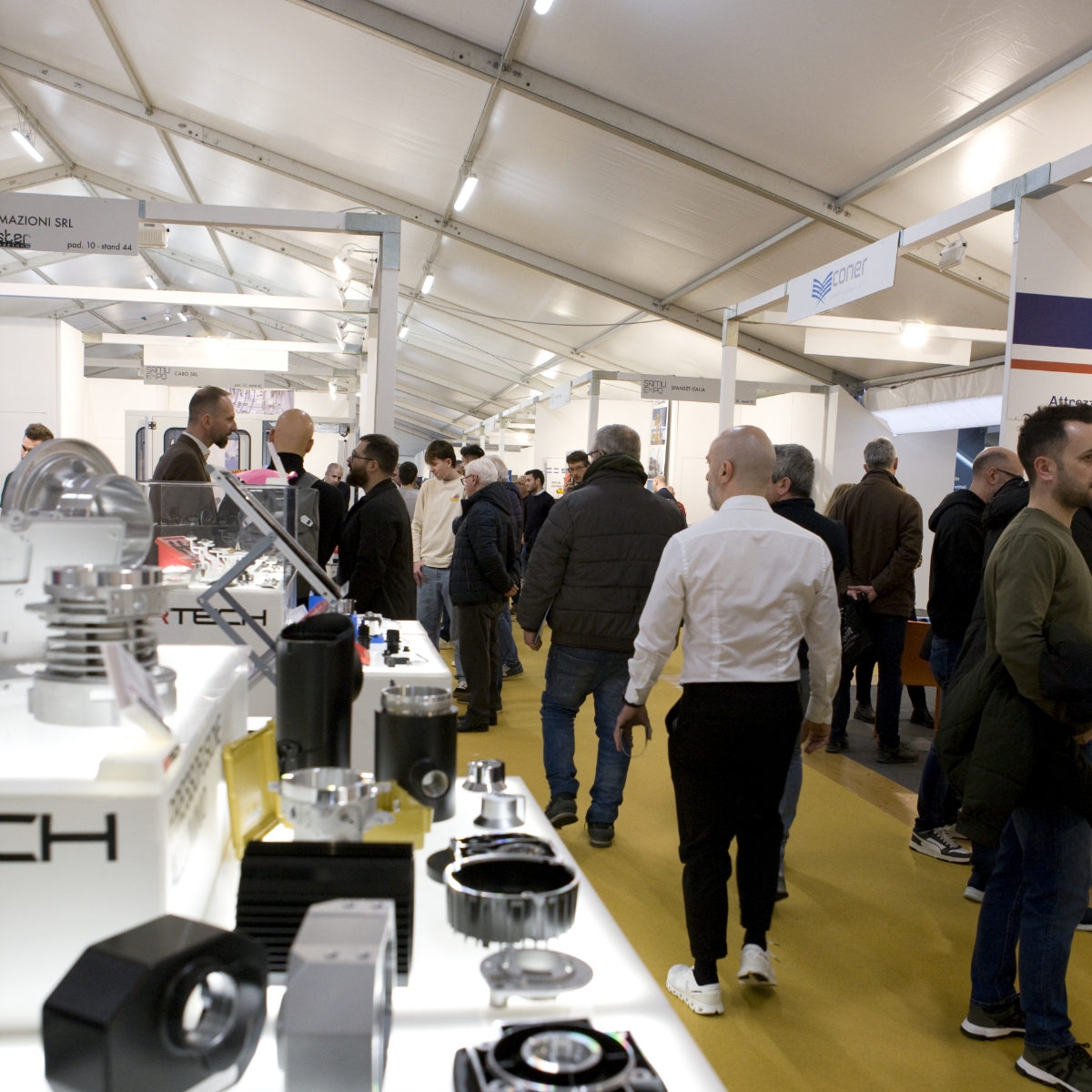 Le persone camminano e parlano all'interno di una sala espositiva affollata, con attrezzature tecniche esposte su tavoli in primo piano e vari stand aziendali allineati ai lati. La sala ha un soffitto a tenda bianco e un corridoio con moquette gialla.