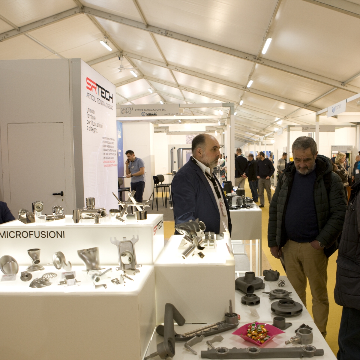 Diversi uomini sono in piedi e parlano in uno stand fieristico che espone parti industriali in metallo sotto luci luminose, con altri stand e partecipanti visibili sullo sfondo all'interno di un grande spazio tendato.