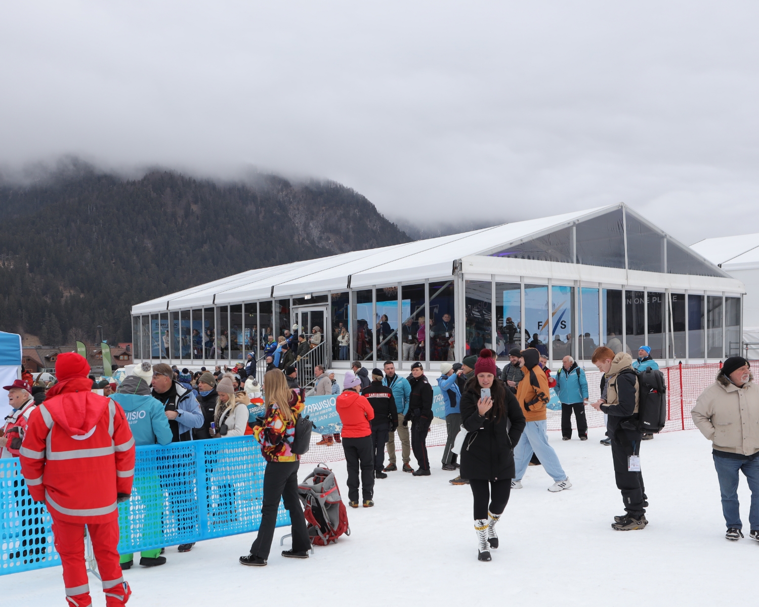 Persone in piedi e a piedi sulla neve vicino a una grande tenda di vetro con molti spettatori all'interno. Sullo sfondo il terreno innevato, le montagne boscose e il cielo nuvoloso. Alcune persone indossano abiti invernali e giacche rosse. È visibile una recinzione blu.