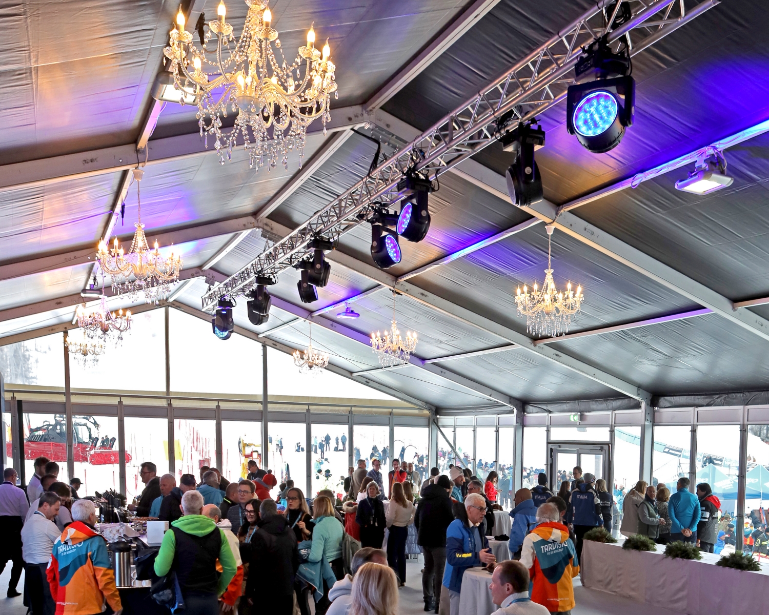 Una grande folla si riunisce all'interno di una tenda spaziosa, con pareti di vetro, lampadari e luci di scena. Le persone si mescolano e parlano, mentre dalle finestre si vedono scene di neve all'aperto con sciatori.