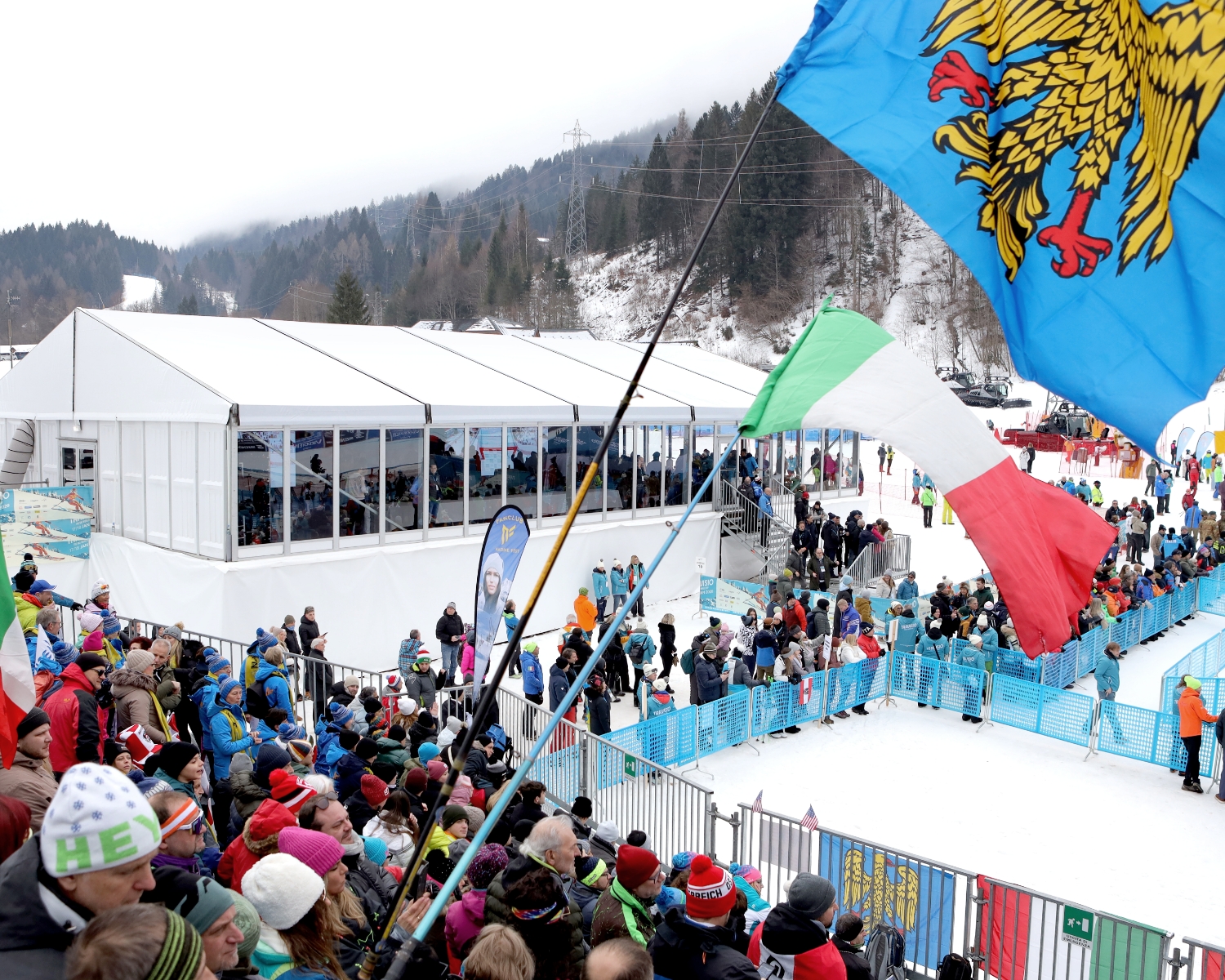 Una folla si raduna a un evento sciistico innevato, sventolando bandiere italiane e regionali vicino a una tenda bianca. Gli spettatori sono vestiti in abiti invernali, con montagne e pini sullo sfondo.