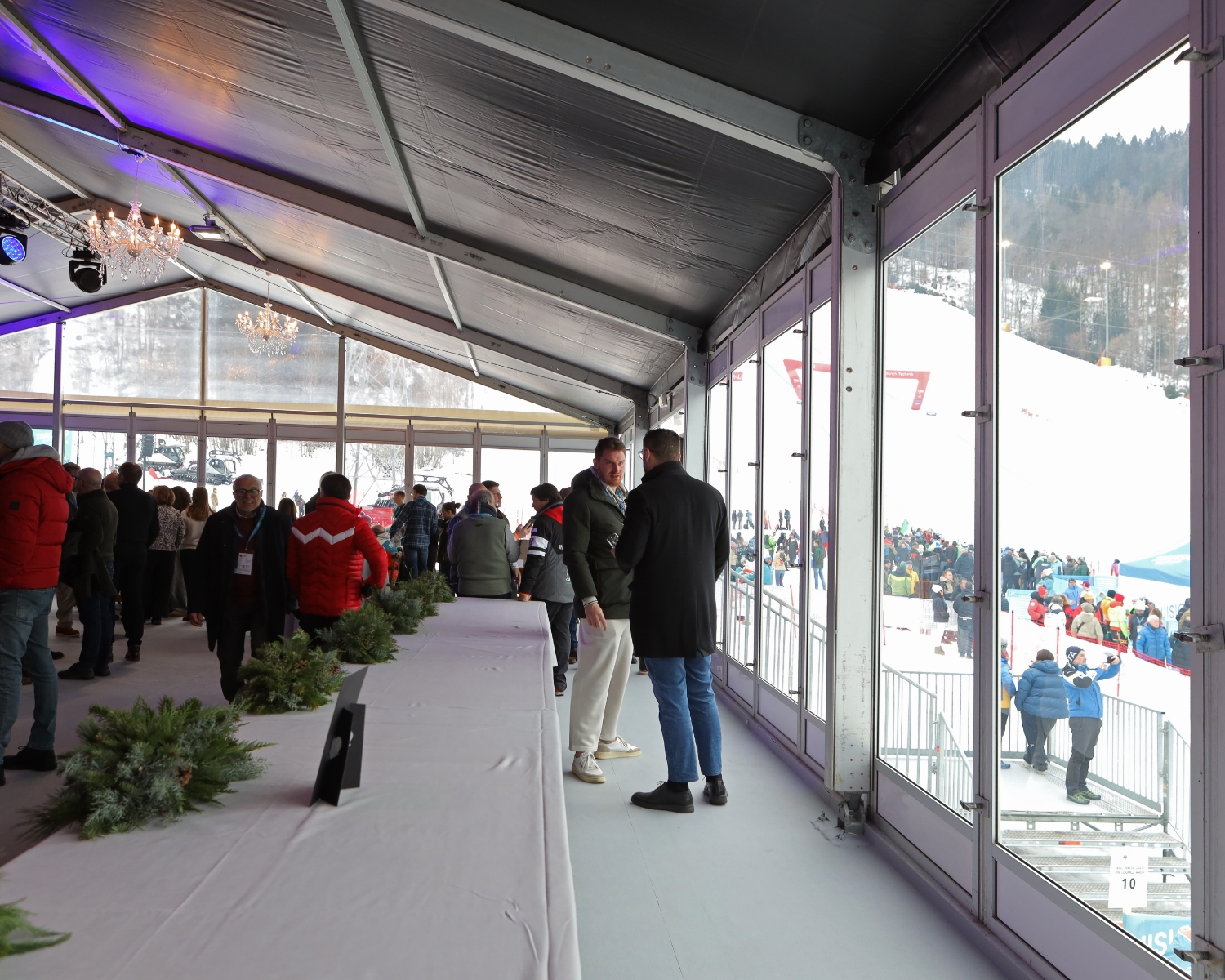 Le persone si trovano all'interno di una tenda dalle pareti di vetro decorate con il verde, che si affaccia su un'area esterna innevata con una pista da sci e spettatori radunati all'esterno. La scena fa pensare a un evento sportivo invernale o a una gara di sci.