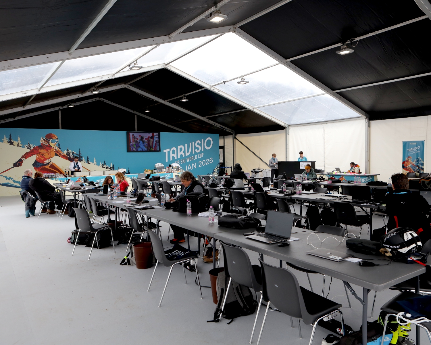 Una grande tenda bianca ospita un centro stampa o media center con molte persone che lavorano ai computer portatili su lunghi tavoli. Sulla parete di fondo si trova il murale di uno sciatore e la scritta "Tarvisio 2026". Lo spazio è ampio e ben illuminato.