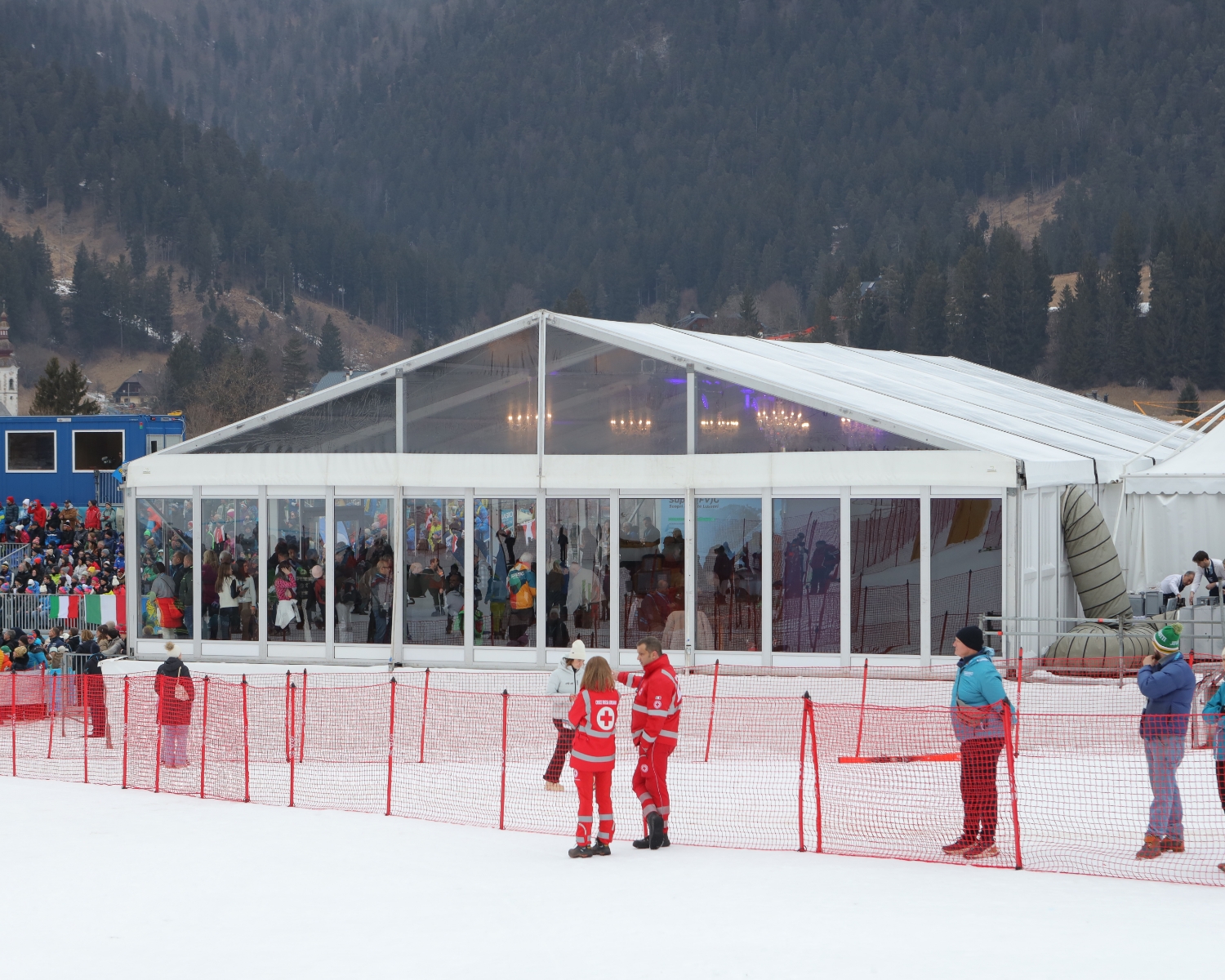 Una grande tenda trasparente si trova in un'area innevata con molte persone all'interno. Due persone in uniforme rossa si trovano vicino a una rete rossa, mentre altri osservano da dietro la rete. Sullo sfondo sono visibili montagne boscose.