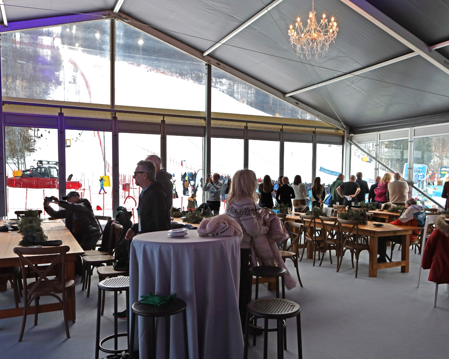 In una grande sala tendata con tavoli e sedie, le persone guardano una pista da sci innevata attraverso grandi finestre; alcuni sono in piedi, altri seduti, e un lampadario pende dal soffitto.