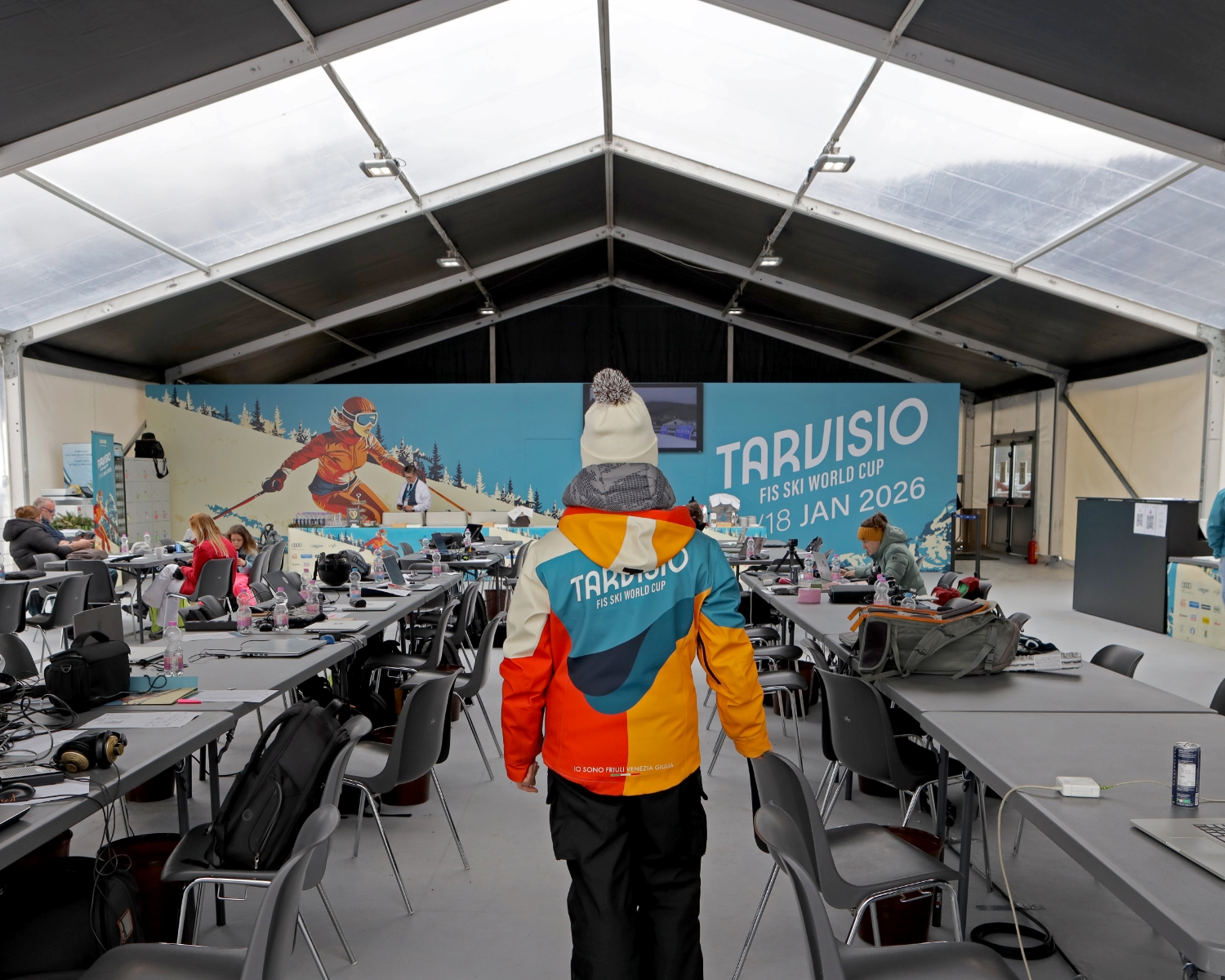 Una persona con una giacca colorata "Tarvisio" si trova all'interno di una grande tenda di lavoro piena di tavoli, sedie e attrezzature. Un murale pubblicizza "Tarvisio FIS Ski World Cup 17-18 gennaio 2026" con uno sciatore sullo sfondo.