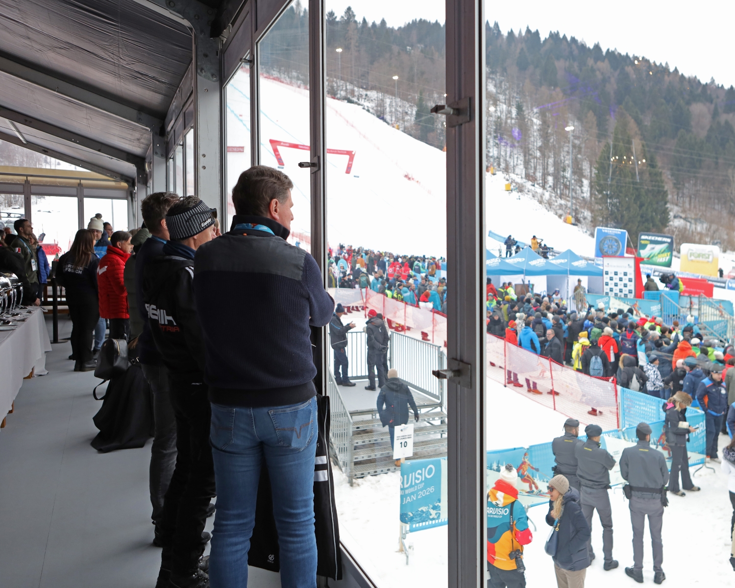 Persone all'interno di un'area chiusa da un vetro assistono a un affollato evento sciistico all'aperto su una montagna innevata. Gli spettatori si riuniscono vicino alle barriere, con tende e alberi sullo sfondo. Alcuni indossano giacche e cappelli invernali.