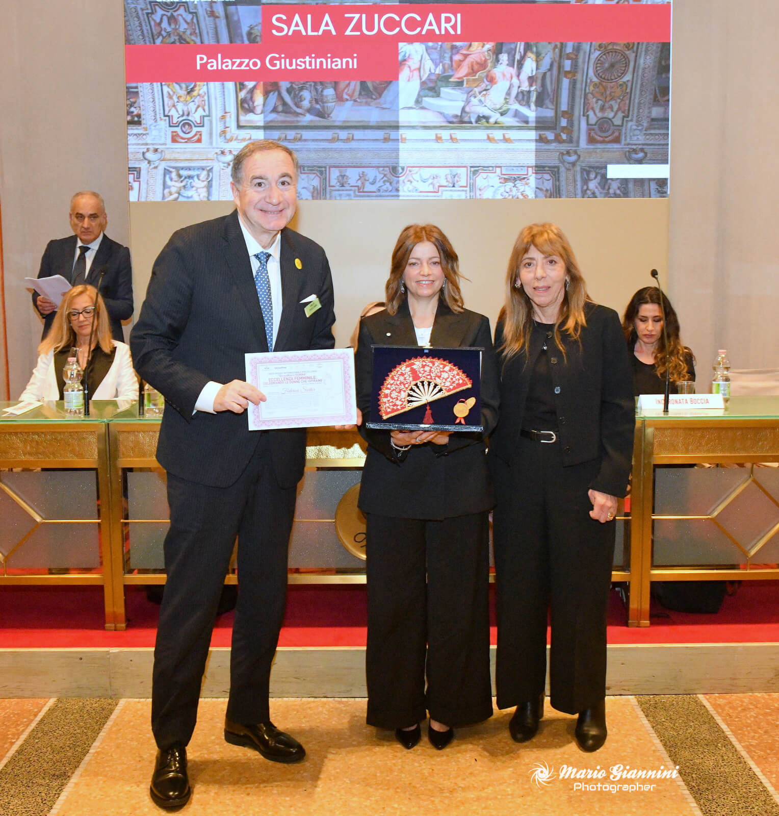 Three people stand smiling at an award ceremony in an ornate room. The person in the center holds a decorative fan and a certificate. A red sign above reads "SALA ZUCCARI Palazzo Giustiniani." Two women and one man are in the foreground.