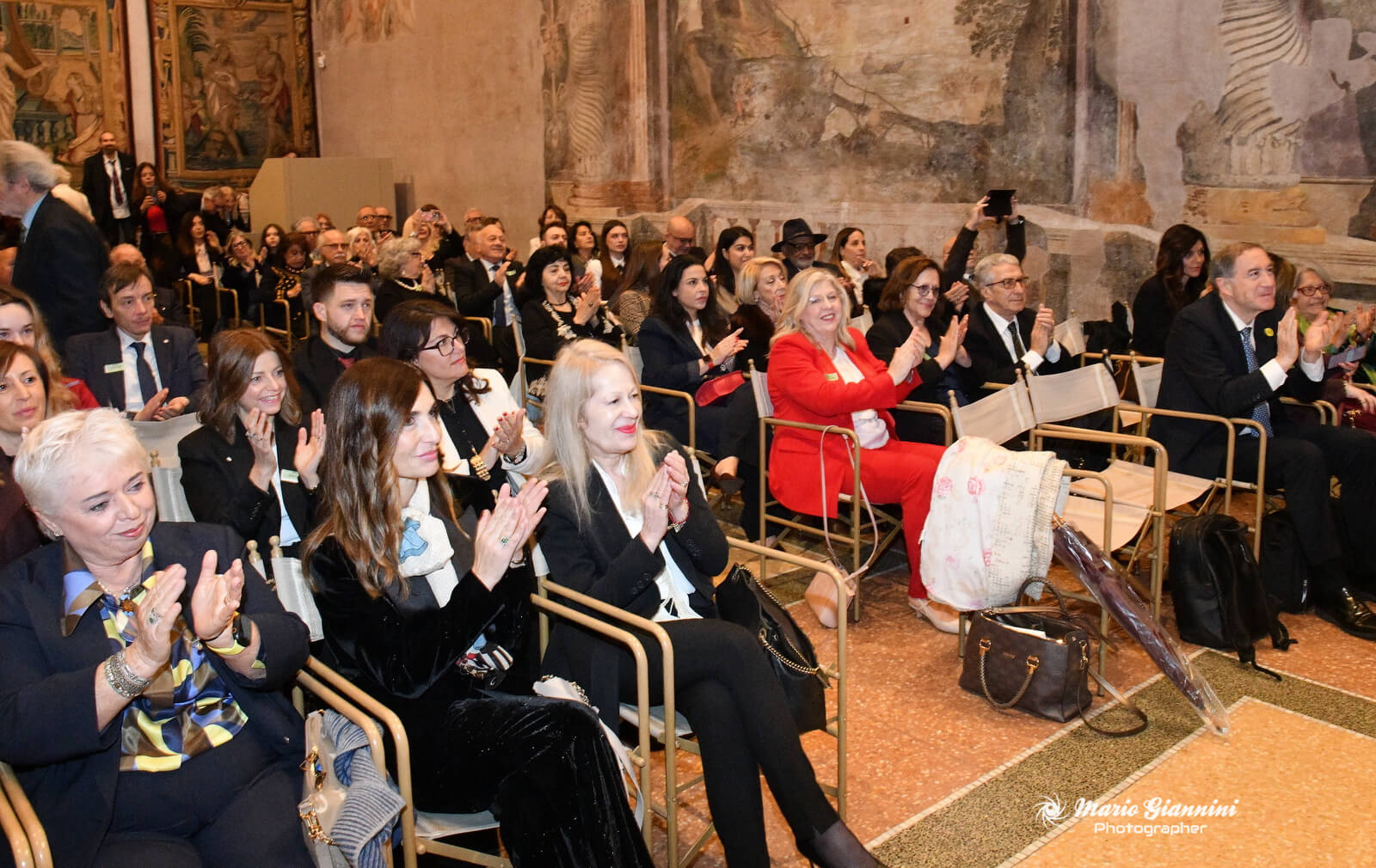 A large group of people, dressed mostly in formal attire, sit in rows of chairs and clap enthusiastically in a grand room with ornate walls and paintings. Some people are smiling and appear to be enjoying an event or ceremony.