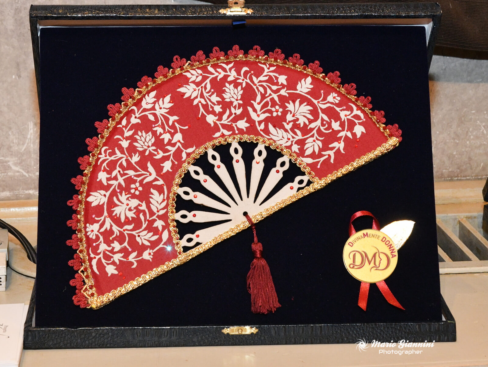 A decorative red and white hand fan with floral patterns and gold trim is displayed in an open black case, alongside a circular ribbon badge marked with "DMD." The photographer's watermark is in the lower right corner.