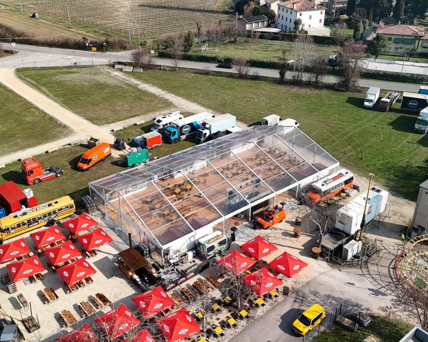 Vista aerea di un evento all'aperto con una grande tenda trasparente, ombrelloni rossi su tavoli da picnic, vari camion e veicoli e campi ed edifici vicini sullo sfondo.