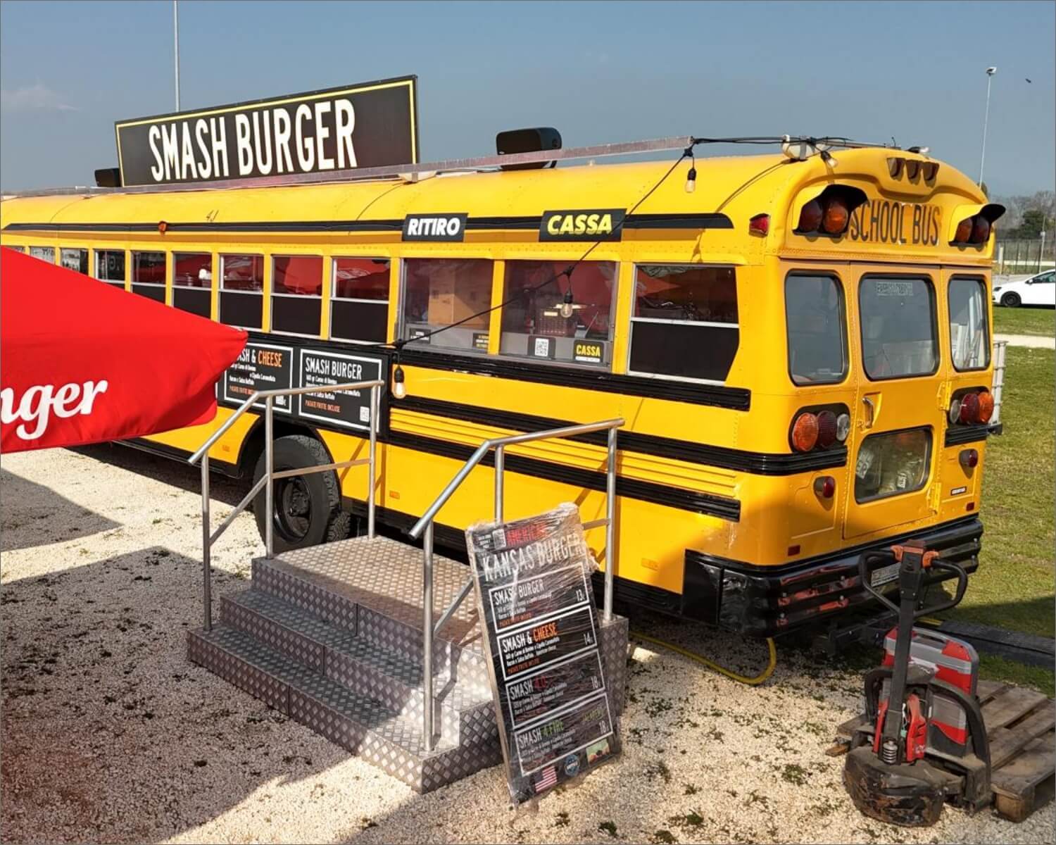 Uno scuolabus giallo trasformato in stand gastronomico si trova all'aperto con un'insegna "Smash Burger" in cima. I gradini conducono all'ingresso e i cartelloni con i menu sono esposti di fronte. Nelle vicinanze sono visibili un ombrellone rosso e un'area erbosa.