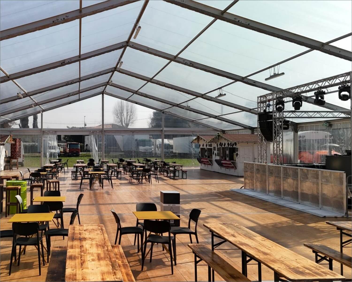 Un'ampia area all'aperto sotto una grande tenda trasparente, con tavoli di legno e sedie nere disposte ordinatamente su un pavimento di legno. Sul lato destro sono allestiti il palco e le luci.