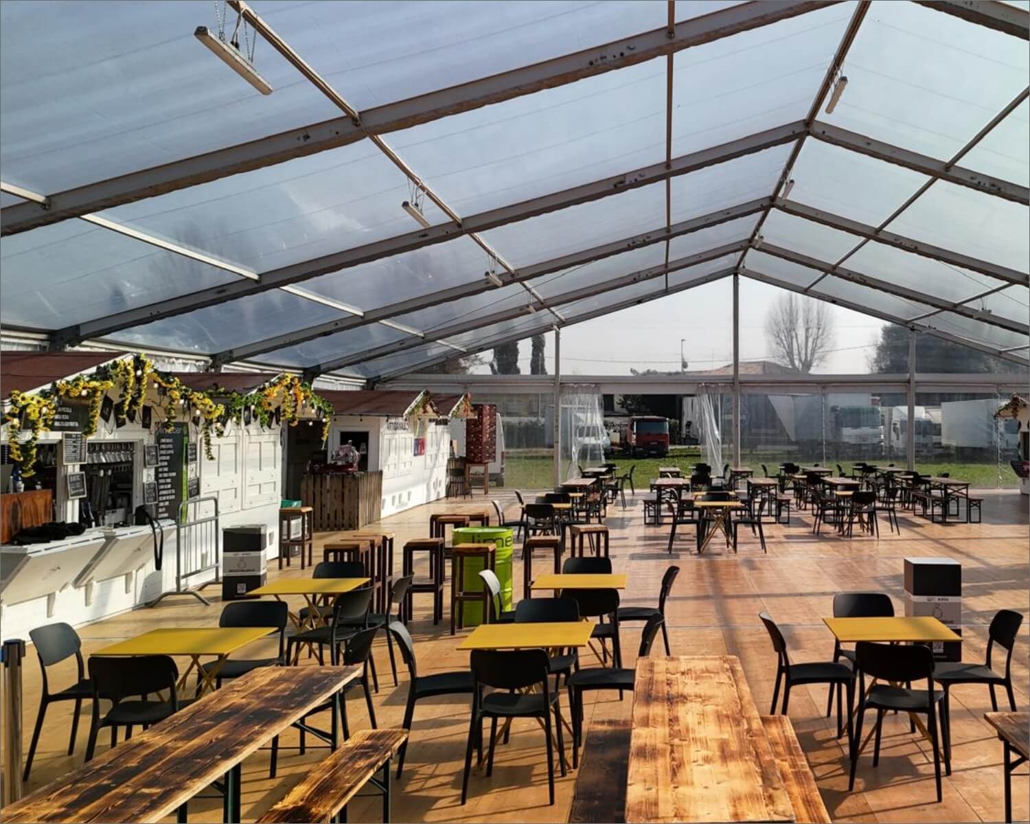 Un ampio spazio all'aperto con tavoli gialli e in legno e sedie nere sotto una grande tenda trasparente, con un bancone decorato sulla sinistra e la luce naturale che entra.