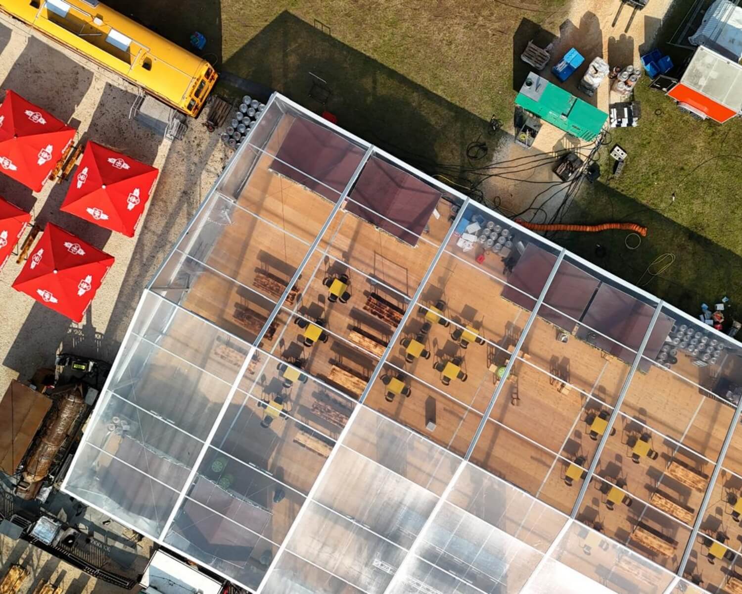 Veduta aerea di una grande tenda trasparente con tavoli e sedie disposti all'interno. Nelle vicinanze ci sono ombrelloni rossi, un autobus giallo, aree erbose e varie attrezzature e strutture.