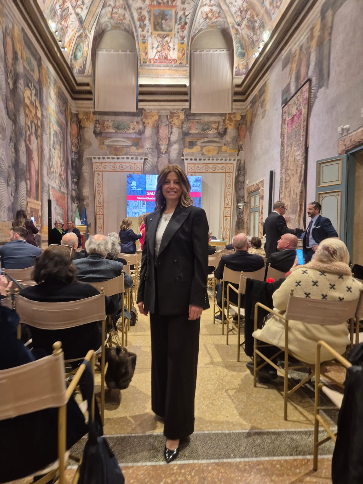 Una donna in abito nero sta in piedi e sorride in una sala ornata con soffitti e pareti dipinte, circondata da persone sedute che partecipano a un evento. Un grande schermo è visibile nella parte anteriore della stanza.