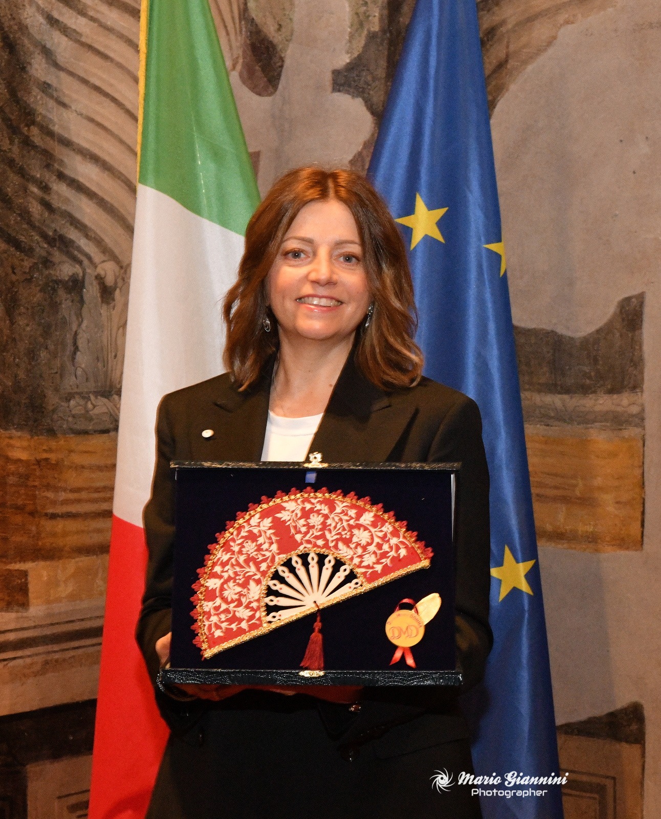 Una donna in blazer nero sorride e tiene in mano un ventaglio ornato di pizzo rosso e bianco in una vetrina. Dietro di lei ci sono le bandiere italiana e dell'Unione Europea e una parete decorativa.