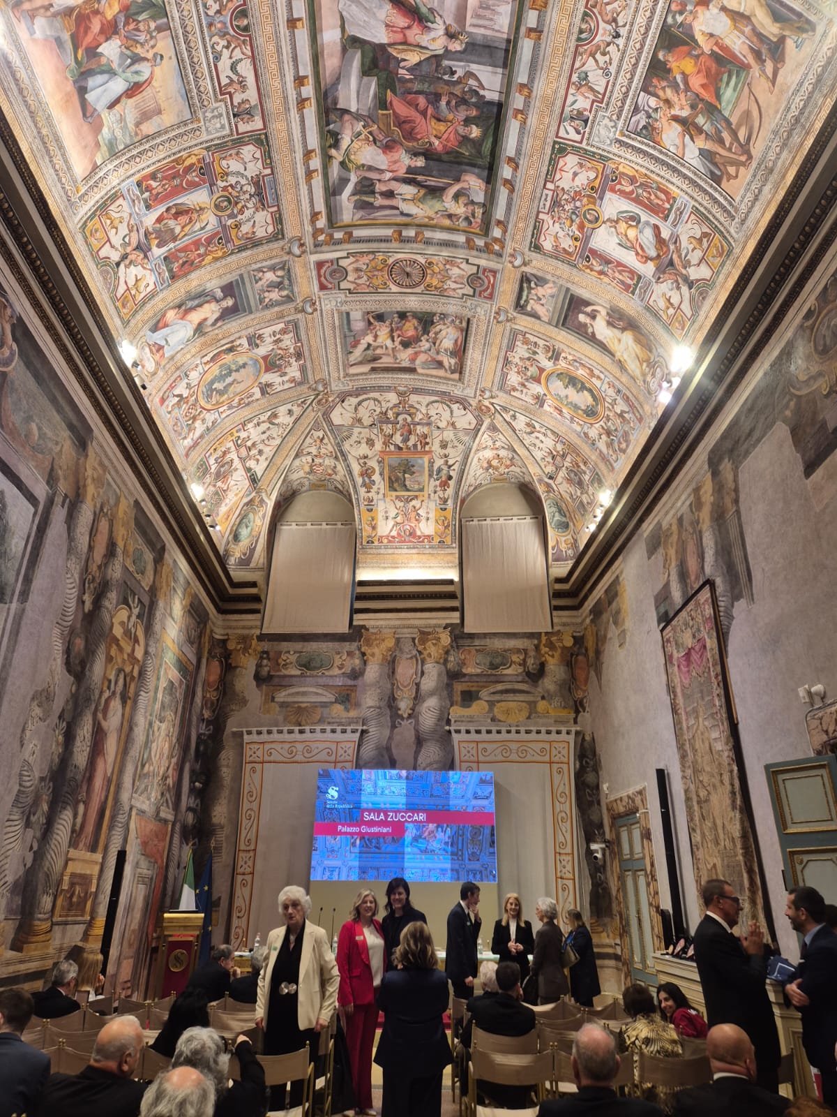 A grand hall with ornate frescoed ceiling and walls, filled with people in formal attire. At the front, a large screen displays "Sala Zuccari." Rows of chairs face the screen, and the atmosphere is elegant and ceremonial.